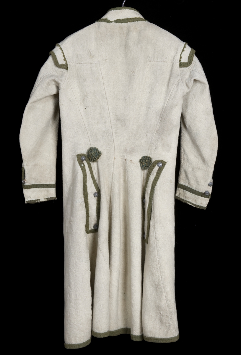 Hvit kjole av ull med grøn dekor av ull.
Midjen bak er markert med to garn dusker.