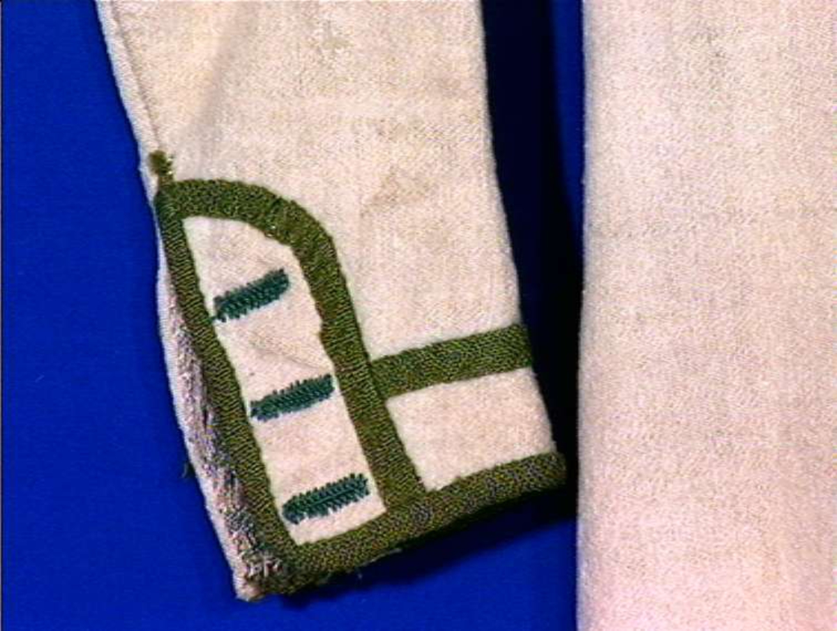 Hvit kjole av ull med grøn dekor av ull.
Midjen bak er markert med to garn dusker.