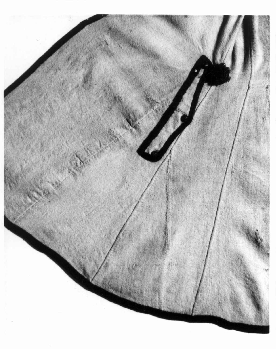 Hvit kjole av ull med grøn dekor av ull.
Midjen bak er markert med to garn dusker.