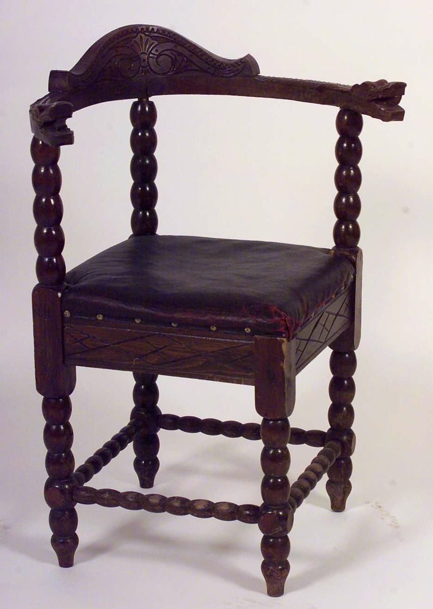 Trestol med diagonalt plassert, firkantet sete med mørkerød voksduk og blomsterformede nagler. Med kuleformer langs bein, sprosser, samt vertikal ryggdel. Øvre del av ryggen er halvsirkelformet og går over til armlenene som har et utskjært dragehode i hver ende.