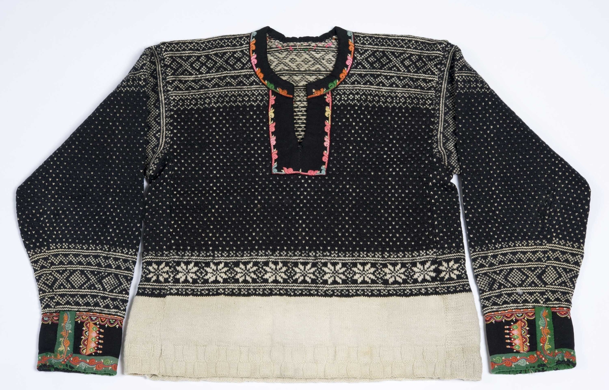 Strikket ullgarn, heimespunnet, svart og hvitt. Broderi i ullgarn. Langs nedrekanten en 13 cm bred hvit bord, resten mønsterstrikket: en bord åttebladroser, en sikksakkbord, en bord kors og kantstilte kvadrat. Langs åpningen foran og halsringingen er påsatt et stykke svart klede, saum i rosa, blått, gult og grønt zephyrgarn. Rundt ermet et 9,3 cm bredt belegg av svart klede, kantet med grønn klede 2,5 cm bred. En brodert bord langs indre og ytre kant av belegget. Aagot Noss 1969. 
