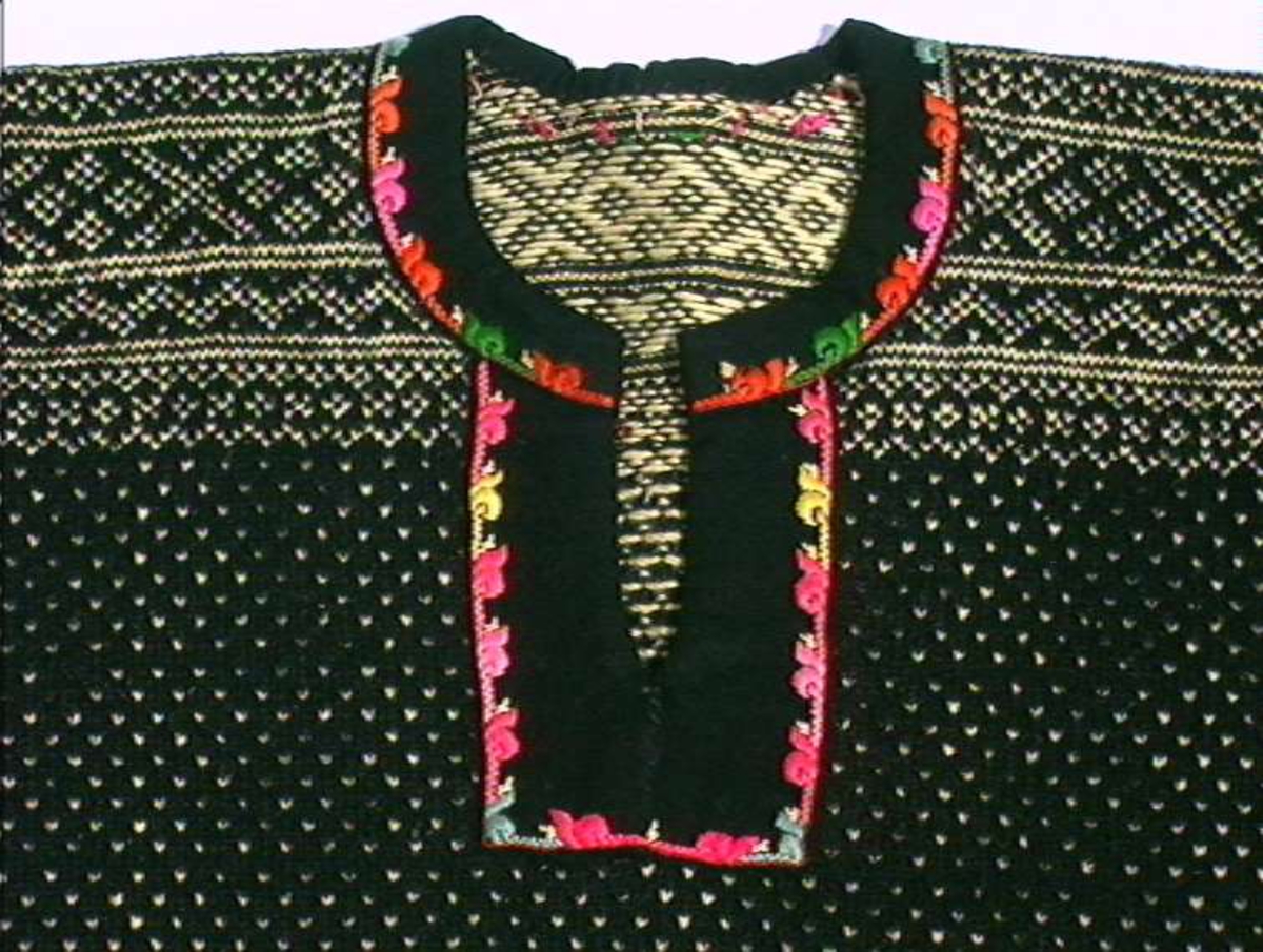 Strikket ullgarn, heimespunnet, svart og hvitt. Broderi i ullgarn. Langs nedrekanten en 13 cm bred hvit bord, resten mønsterstrikket: en bord åttebladroser, en sikksakkbord, en bord kors og kantstilte kvadrat. Langs åpningen foran og halsringingen er påsatt et stykke svart klede, saum i rosa, blått, gult og grønt zephyrgarn. Rundt ermet et 9,3 cm bredt belegg av svart klede, kantet med grønn klede 2,5 cm bred. En brodert bord langs indre og ytre kant av belegget. Aagot Noss 1969. 
