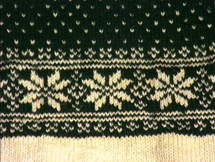 Strikket ullgarn, heimespunnet, svart og hvitt. Broderi i ullgarn. Langs nedrekanten en 13 cm bred hvit bord, resten mønsterstrikket: en bord åttebladroser, en sikksakkbord, en bord kors og kantstilte kvadrat. Langs åpningen foran og halsringingen er påsatt et stykke svart klede, saum i rosa, blått, gult og grønt zephyrgarn. Rundt ermet et 9,3 cm bredt belegg av svart klede, kantet med grønn klede 2,5 cm bred. En brodert bord langs indre og ytre kant av belegget. Aagot Noss 1969. 

