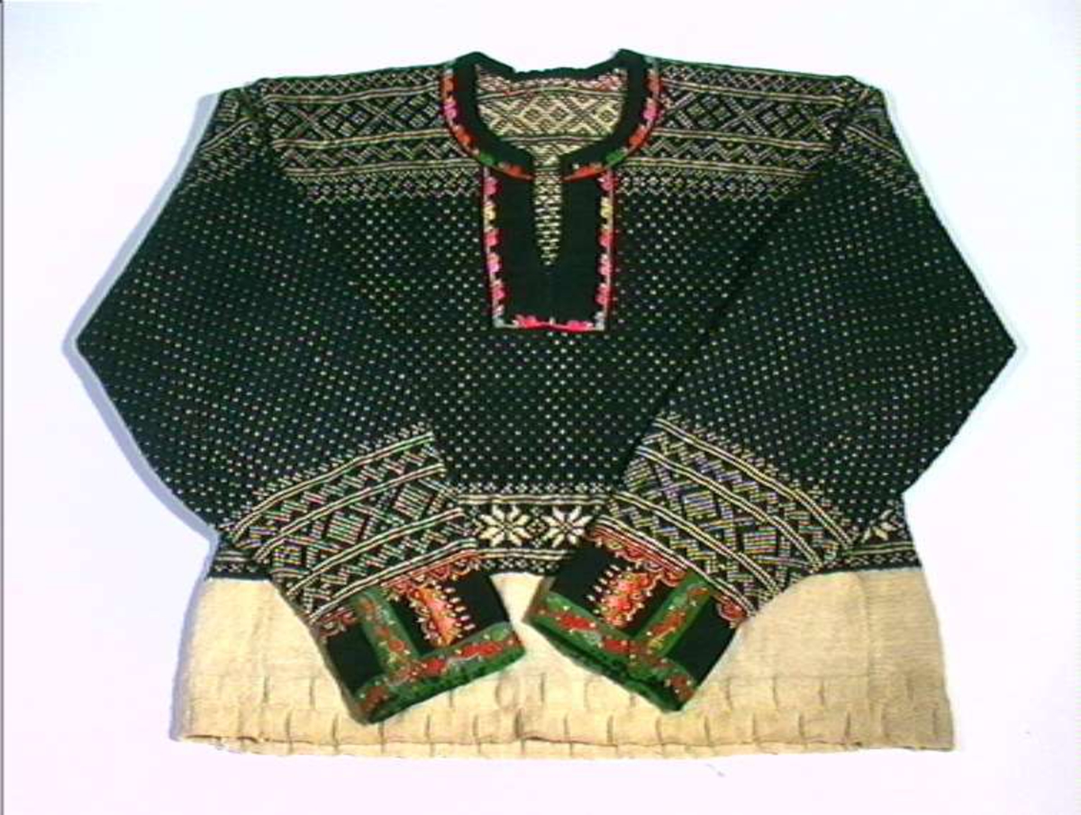 Strikket ullgarn, heimespunnet, svart og hvitt. Broderi i ullgarn. Langs nedrekanten en 13 cm bred hvit bord, resten mønsterstrikket: en bord åttebladroser, en sikksakkbord, en bord kors og kantstilte kvadrat. Langs åpningen foran og halsringingen er påsatt et stykke svart klede, saum i rosa, blått, gult og grønt zephyrgarn. Rundt ermet et 9,3 cm bredt belegg av svart klede, kantet med grønn klede 2,5 cm bred. En brodert bord langs indre og ytre kant av belegget. Aagot Noss 1969. 
