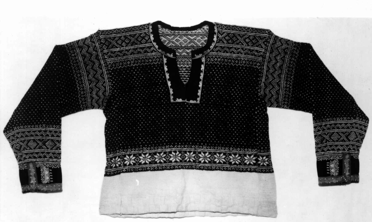 Strikket ullgarn, heimespunnet, svart og hvitt. Broderi i ullgarn. Langs nedrekanten en 13 cm bred hvit bord, resten mønsterstrikket: en bord åttebladroser, en sikksakkbord, en bord kors og kantstilte kvadrat. Langs åpningen foran og halsringingen er påsatt et stykke svart klede, saum i rosa, blått, gult og grønt zephyrgarn. Rundt ermet et 9,3 cm bredt belegg av svart klede, kantet med grønn klede 2,5 cm bred. En brodert bord langs indre og ytre kant av belegget. Aagot Noss 1969. 
