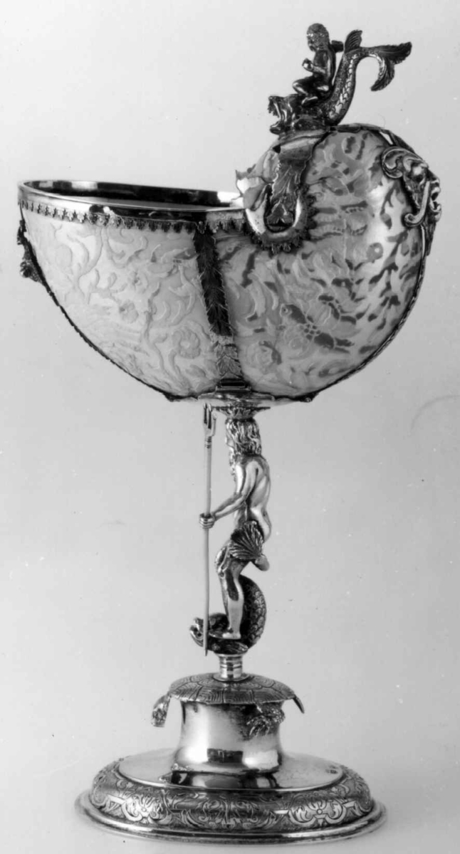 Skall av blekksprutarten Nautilus. Skåret dekorasjon, fot og innfatning sølv, støpt og gravert dekorasjon. Stpl: MF/H//1720 Morten Finchenhagen, Christiania. Skallet er brunflammet i overflaten. Nautilusblekkspruten lever i sydlige hav omkring Filippinene.