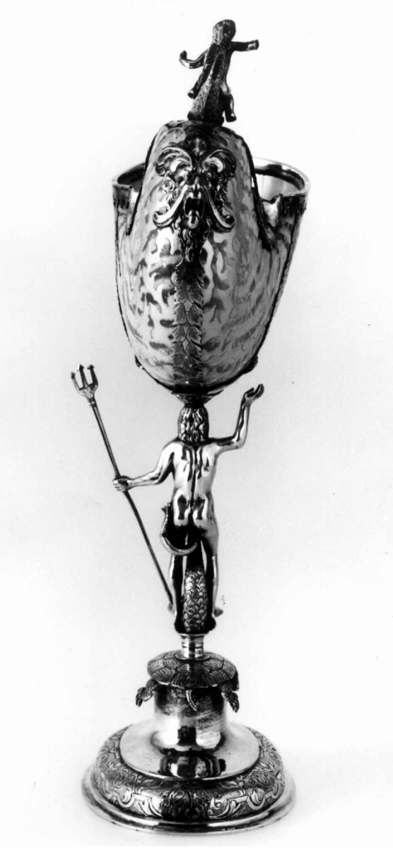 Skall av blekksprutarten Nautilus. Skåret dekorasjon, fot og innfatning sølv, støpt og gravert dekorasjon. Stpl: MF/H//1720 Morten Finchenhagen, Christiania. Skallet er brunflammet i overflaten. Nautilusblekkspruten lever i sydlige hav omkring Filippinene.