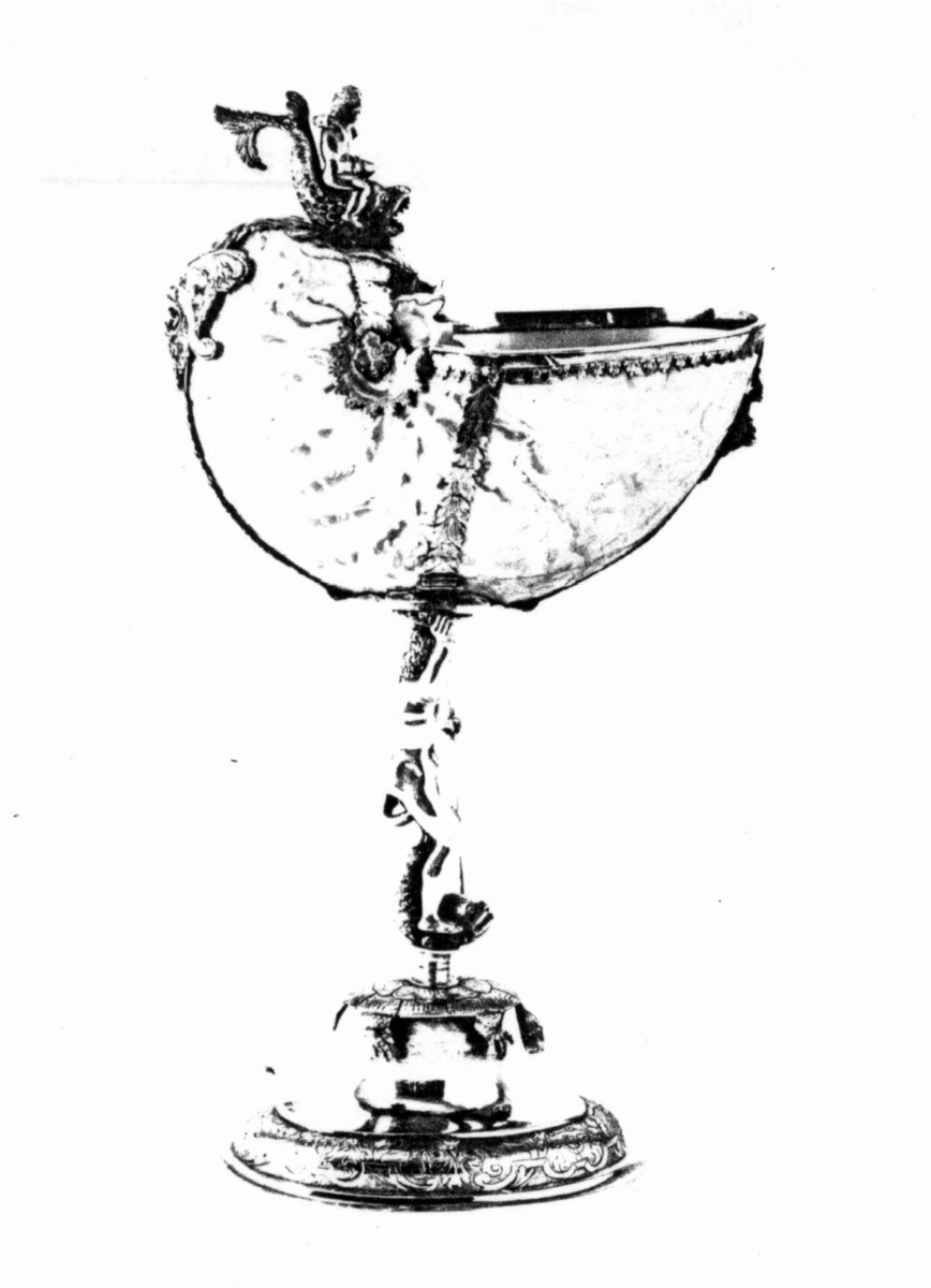 Skall av blekksprutarten Nautilus. Skåret dekorasjon, fot og innfatning sølv, støpt og gravert dekorasjon. Stpl: MF/H//1720 Morten Finchenhagen, Christiania. Skallet er brunflammet i overflaten. Nautilusblekkspruten lever i sydlige hav omkring Filippinene.