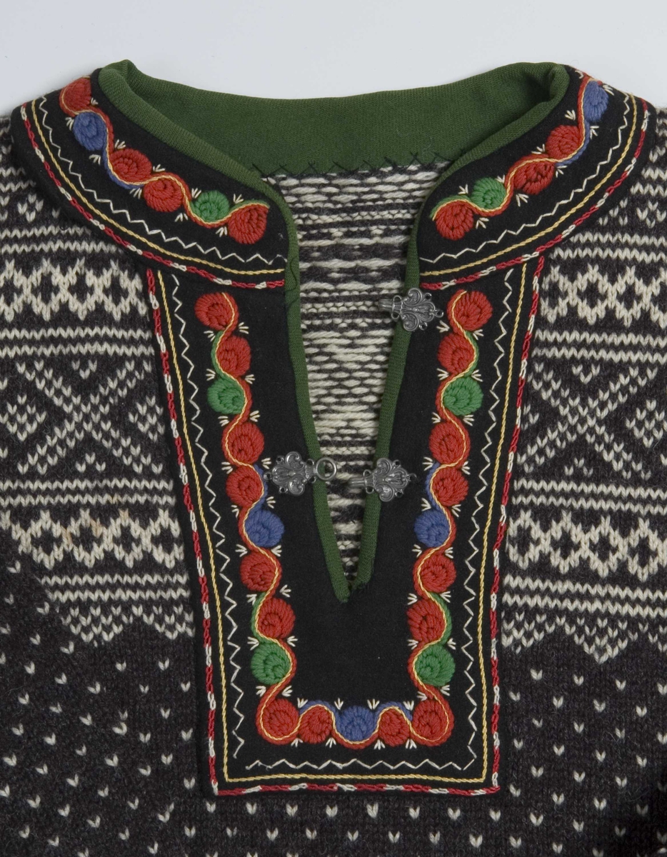 Grå kofte i genserfasong. Splitt i halsen med 2 par metallhekter. Hvitt Setesdalsmønster og brodert kant langs halslinning og splitt. 