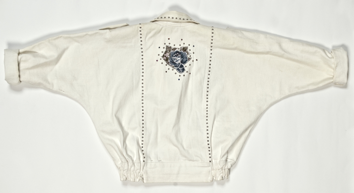Hvit jakke i bomull med dekor av kuler og fargestener med applikerte felt av blomstermønstret tekstil med glitter på. Tilhørende shorts med samme farger og dekor. 
