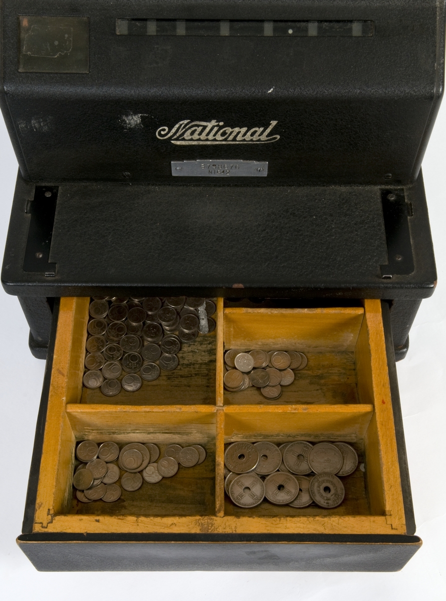 Sort kassaapparat med pengeskuff og sveiv. Maks innslag 99 kroner og 99 øre.