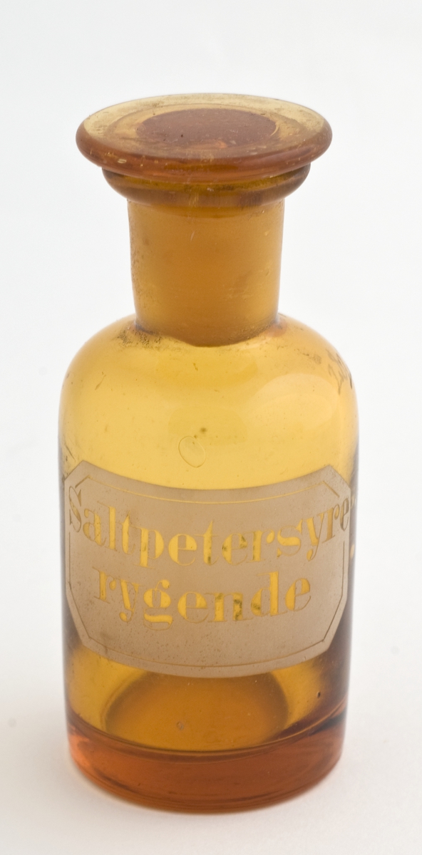 Brunt standglass med rund propp. Hvit etikett med utspart skrift, rektangulær med avkuttede hjørner.