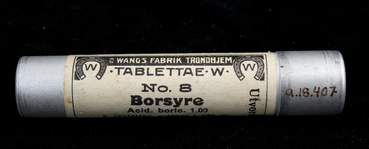 Tablettrør av metall med skrukork og innhold. Hvit etikett med sort trykk.