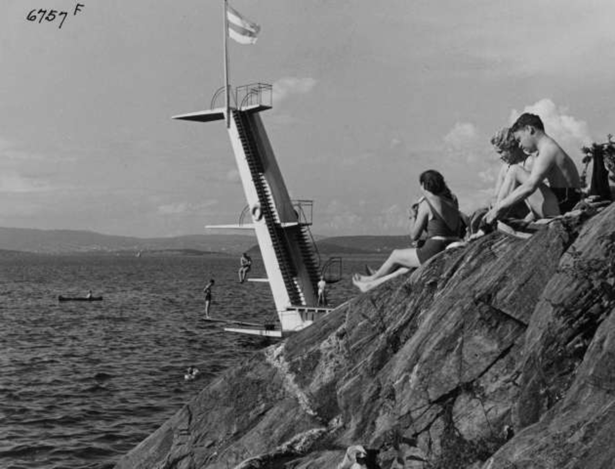 Strandliv med badegjester som soler seg på svaberget ved sjøen på Ingierstrand bad utenfor Oslo. Stupetårn i bakgrunnen.