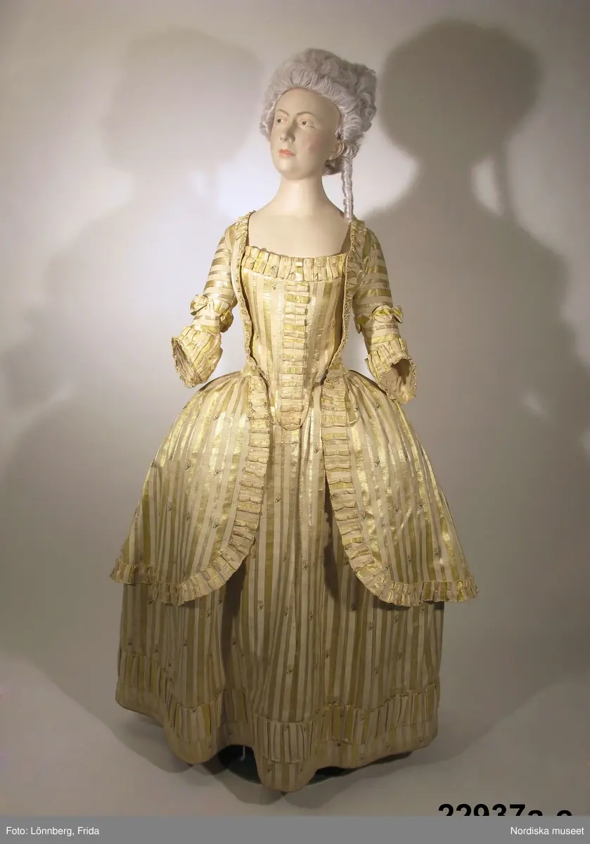 Klänning i 3 delar, robe a, kjol b samt bröstlapp c. 
Klänningen sydd i snittet för kvinnodräkt i Gustaf III:s nationella dräkt.
Av halvsiden med varp av naturvitt silke och tjockare inslag i gulvitt bomullsgarn, vilket ger viss ripseffekt. Ränder i gult silke med rosa konturer i inslagssatin,  mellan ränderna spridda små blommor i rosa och grönt silke broderade i tambursöm.
a. Robe avskuren fram, helskuren i ryggen. 2 framstycken, 4 sidstycken, 2 ryggstycken med veck bak från midjan, rveckad kjoldel mot fram-och sidstycken med avrundade framkanter. Isydd armbågslång snäv ärm i ett stycke, kanten svängd för ambågen. Kring ärmkant och runt hela roben veckade bandkantningar av klänningens tyg, på ärmen även rynkvolang av tyget strax ovanför kanten. Foder av oblekt linnelärft i livet. Ett knytband i midjan.
b. kjol i ett stycke med lagda veck i midjan mot smalt linningsband av rostrött linne, sprund i båda sidor. En bit från nederkanten veckbandsgarnering av samma slag som på roben. Kjolen infållad med 7 cm bred remsa av roströd linnelärft, som syns som en smal passepoal i nederkanten. Rektangulär lapp påsydd nedanför midjan
Anm. Färgen tämligen blekt, de gula sidenbårderna har varit klargula och den rosa färgen tydligare, vilket syns på undanskymda ställen. 
c. Bröstlapp i klänningens tyg. Fyrkantigt stycke som svänger ned i en snibb, Veckbandsgarnering som på roben mitt fram och längs överkanten. Foder av linnelärft. Ett knytband nedtill i var sida.
Anm. På bröstlappens insida 3 stålfjädrar fastsydda med vita bomullsband som är sekundära, det ena knytbandet  är också ersatt med ett nyare. Troligen har bröstlappen inte haft någon styvnad från början.
Se Eva Bergman: Nationella dräkten, Nordiska museet 193?  sid
/Berit Eldvik 2009-04-27