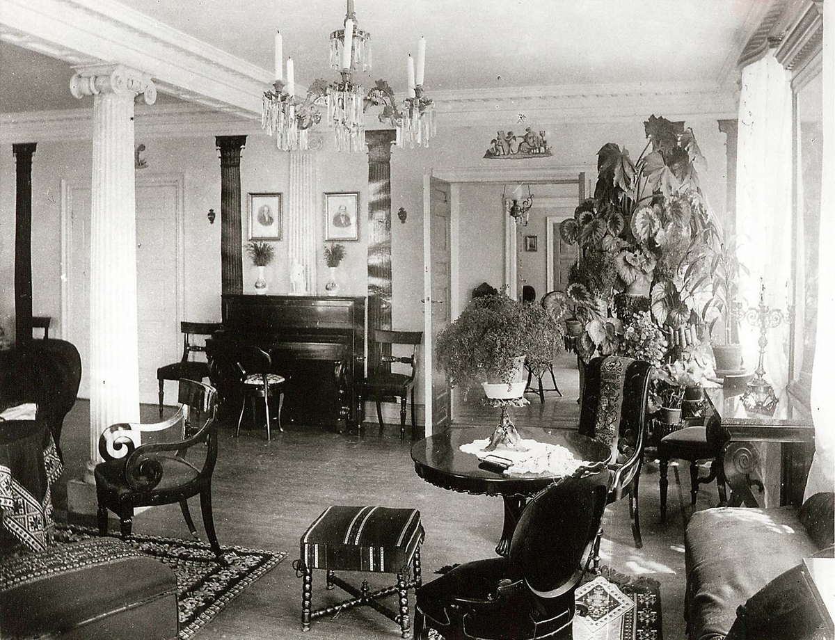 Strømsbo gård, Strømsbu, Arendal. Interiør, salen. Søyler, div. møbler som gyngestol, potteplanter, piano. Krystall lysekrone i taket. H.del av panorama, se NF.04551-015. Hovedbygningen er oppført 1760/61, men fikk sitt nåværende preg i 1804 da den ble overtatt av statshauptmann Peter Herlofson. Fredet.
Fra serie (NF.04551-001 til -021) fra billedmappen "Strømsbo", gård på Strømsbu i Arendal tilh. familien Herlofson. Bildene er fra ulike eiendommer og steder i Arendal tilknyttet familien, fotografert av P.A. Thorèn, Chra.