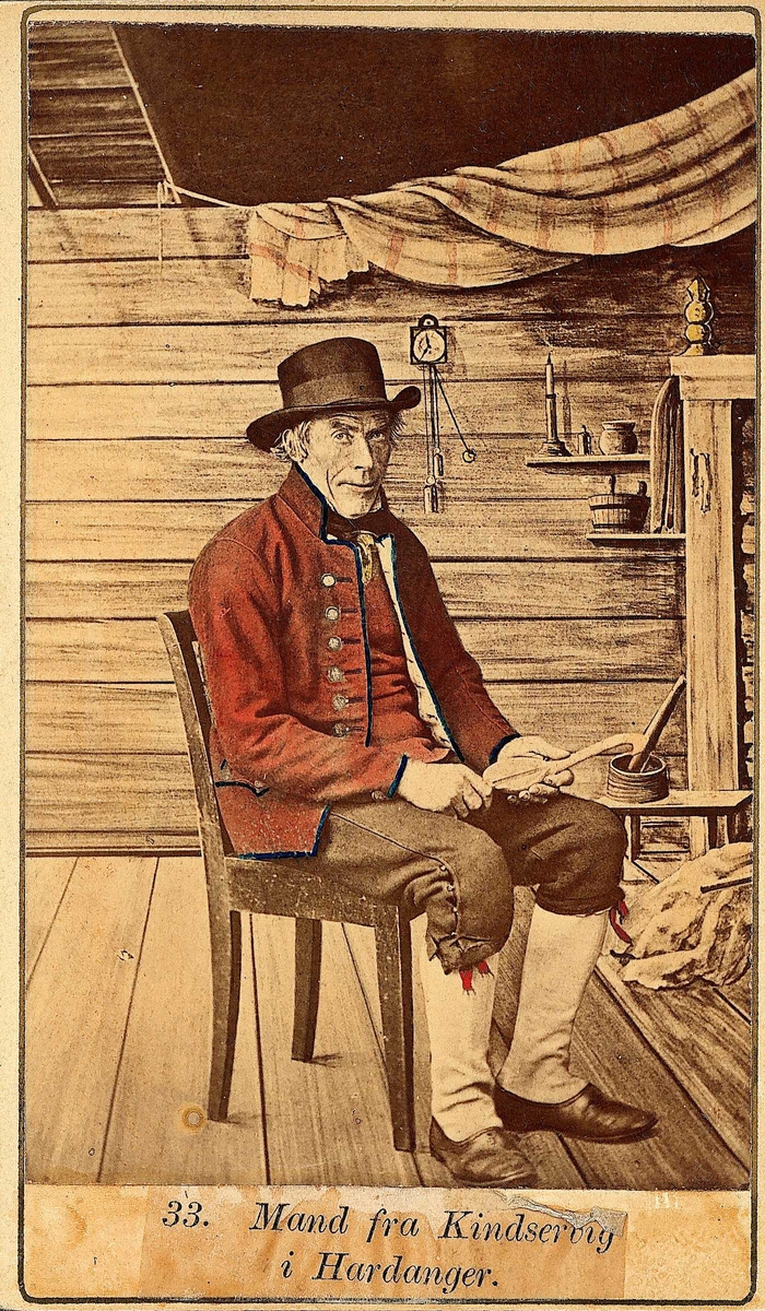 33. Mannsdrakt, Kinsarvik, Ullensvang. Portrett av mann med hatt sittende i fotoatelier med malt stueinteriør som bakgrunn.
Fra serien "Norske Nationaldragter" tatt av fotograf Marcus Selmer (1819-1900), Bergen. 