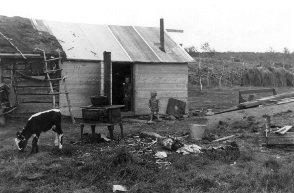 Forsiden av boligen med to barn ved døråpningen, en ovn i forgrunnen. Mikkel Johansen Utsis gård. Hemmujavve 1953.