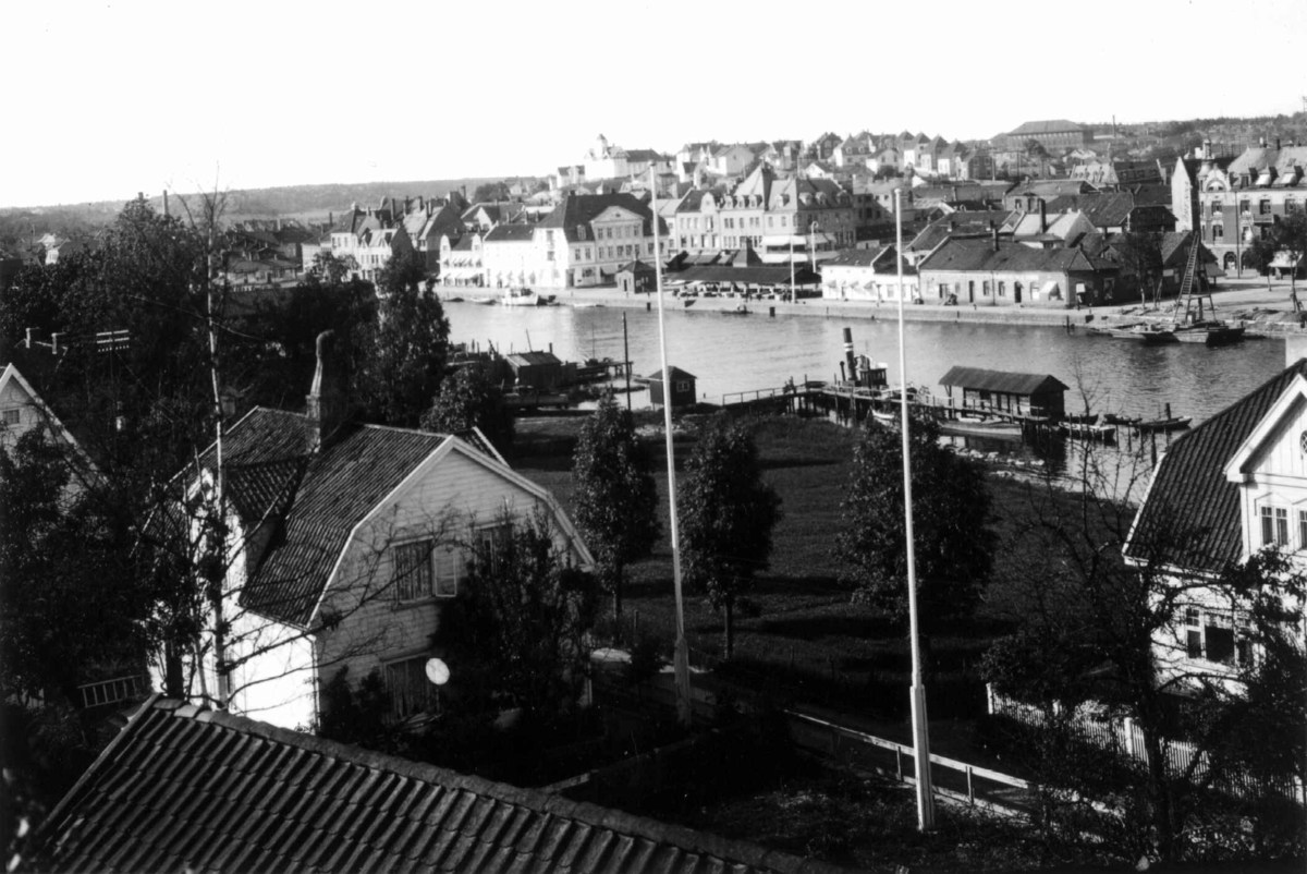 Fredrikstad, Østfold. 1930. Oversiktsbilde over byen.
Villaer i forgrunnen med hager ned til elva. Fortøyde båter langs elva. Bybebyggelse i bakgrunnen.