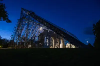 Mørke trær omkranser et trekantet glassbygg over en opplyst kirkeruin, med mørk blå nattehimmel over.