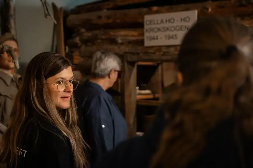 En kvinne ser bakover mot en annen kvinne mens hun forteller. I bakgrunnen ser man en tredje kvinne som ser på en utstilling i Hjemmestyrkemuseet. Man ser også et uskarpt skilt hvor man kan se at det står "Cella Ho-Ho, Krokskogen, 1944-1945" 