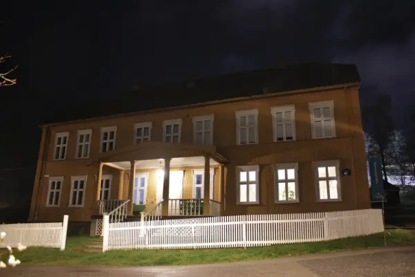 Gult hus i empirestil med hvitt stakittgjerdet. Bildet er tatt om kvelden, og det er lys i vinduet.