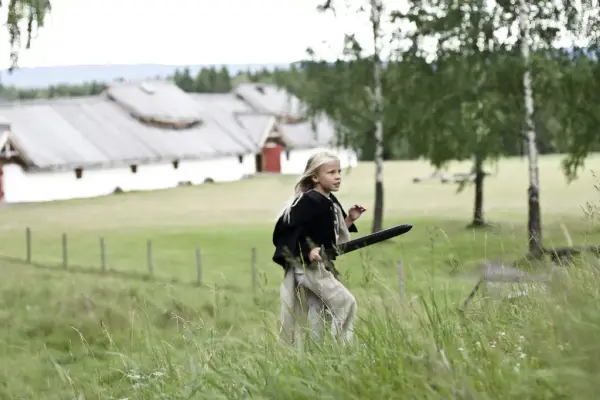 Ung jente løper i gravfeltet med sverd i hånden. I bakgrunnen vises langhuset