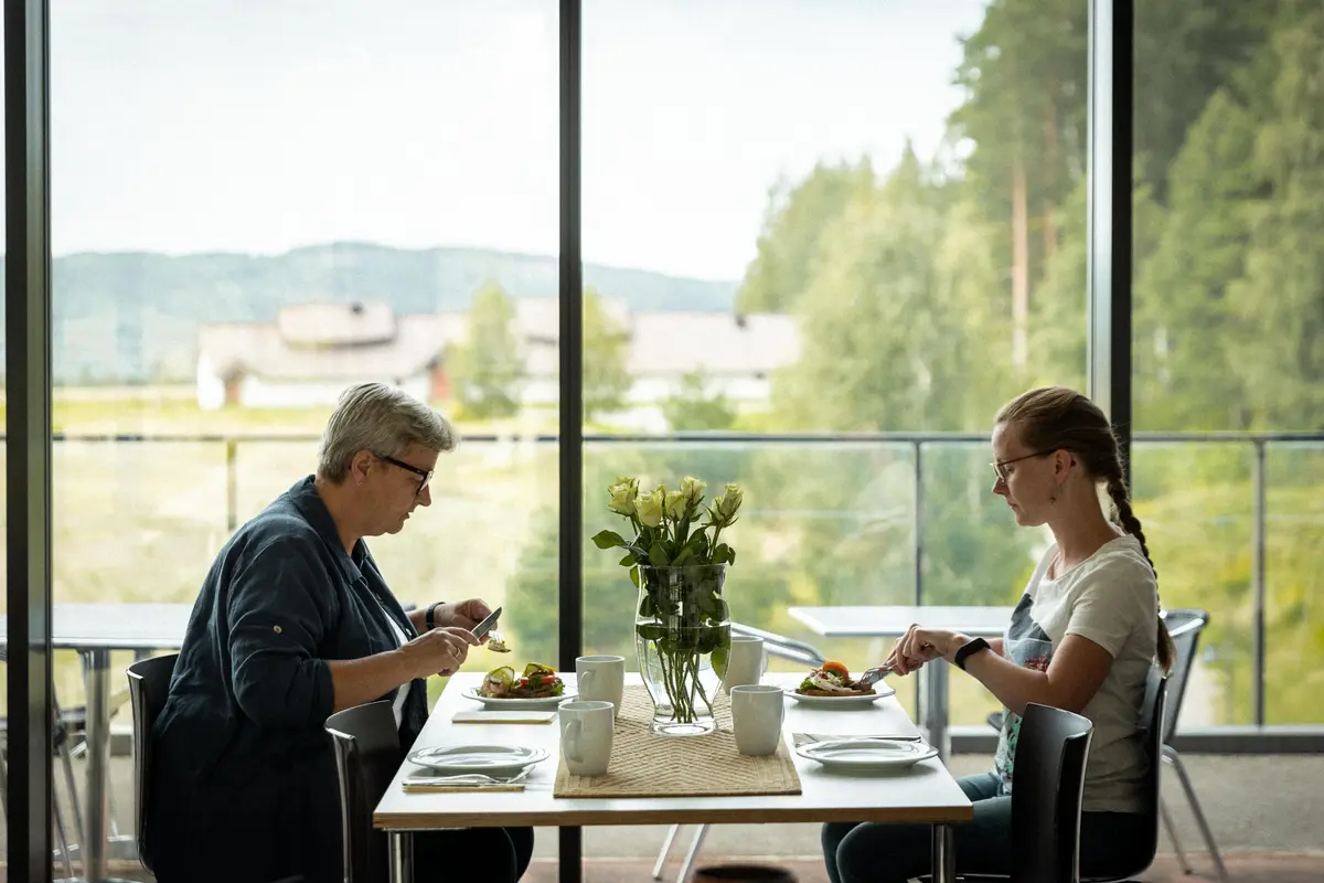 To voksne damer spiser lunsj i museumsbygningen med utsikt til langhuset