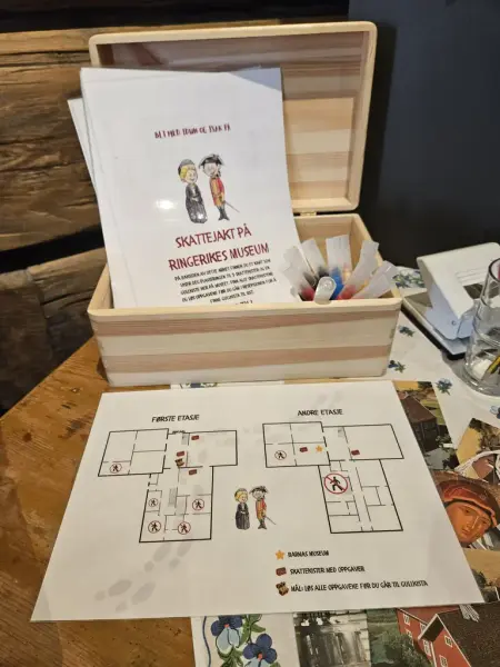 Et bord med en åpen trekiste som inneholder instruksjoner for en skattejakt-aktivitet, kart og blyanter. 