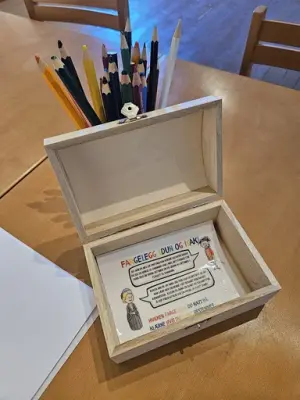 Et bord med en åpen trekiste som inneholder en tegne-oppgave. Rundt står fargeblyanter, som gir inntrykk av en interaktiv eller kreativ aktivitet.