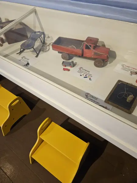 En glassmonter med små leker, som en rød lastebil og andre små gjenstander. Under monteren står gule krakker som viser at utstillingen er tilpasset barn.