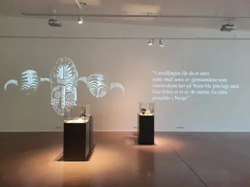 I utstilling er det tekst på vegg: "I utstillingen får du et nært møte med noen av gjenstandene som menneskene her på Veien ble gravlagt med. Gravfeltet er et av de største fra eldre jernalder i Norge" På gulvet står det to montre med gjenstander i.