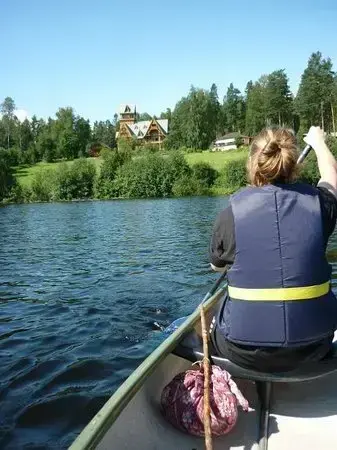 En kvinne padler i kano mot land. I bakgrunnen er Bjørøya, med gressplen og Villa Fridheim. Bildet er tatt fra kanoen.