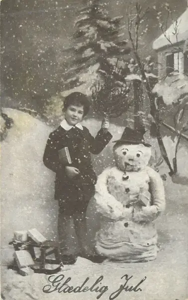 En gutt og en snømann i et snødekket landskap og teksten "Glædelig jul"