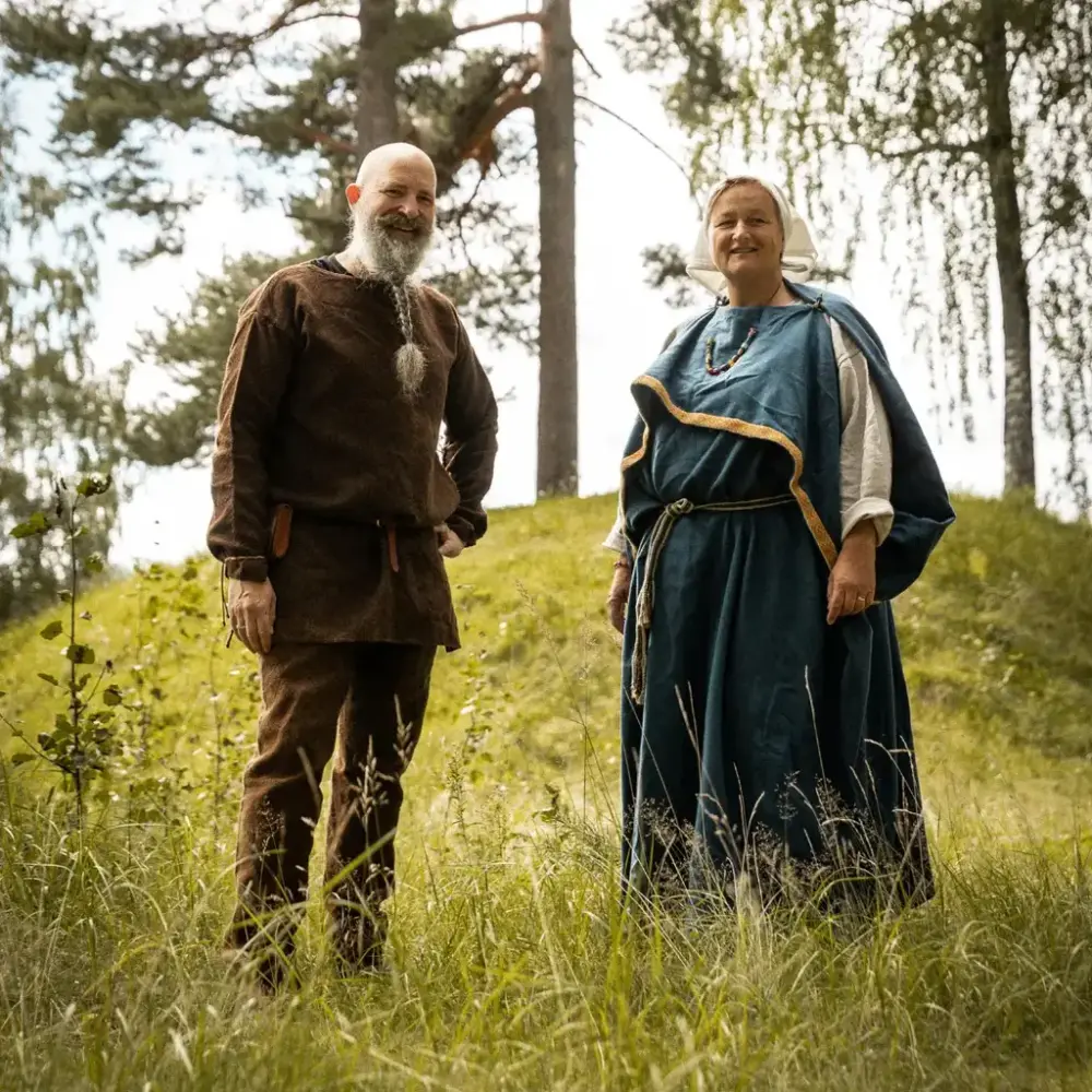 Mann i brun jernalderdrakt og kvinne i blå jernalderkjole står i gresset foran en gravhaug