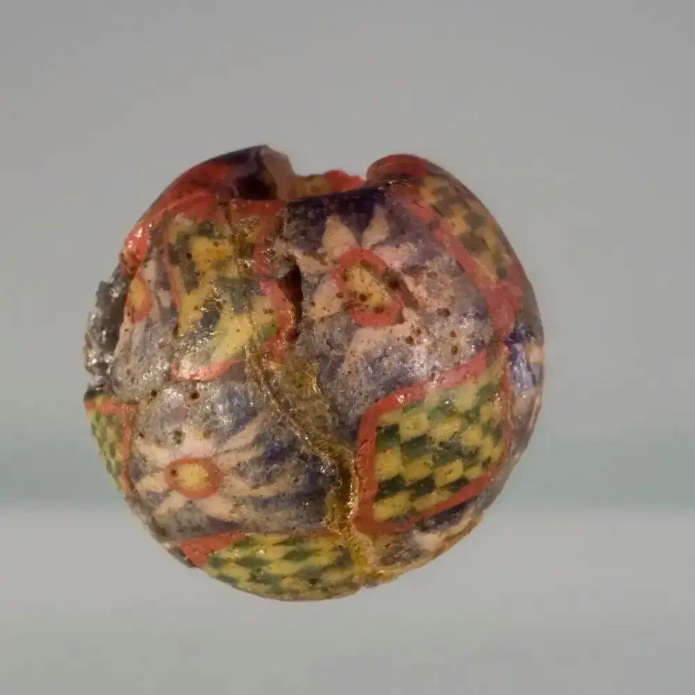 Mosaikkperle med fargene blå, rød hvit, gul og grønn. Noe sjakkmønster i fargene gult og grønt. Blomster som er hvite, røde og gule i midten og blå bakgrunn. 