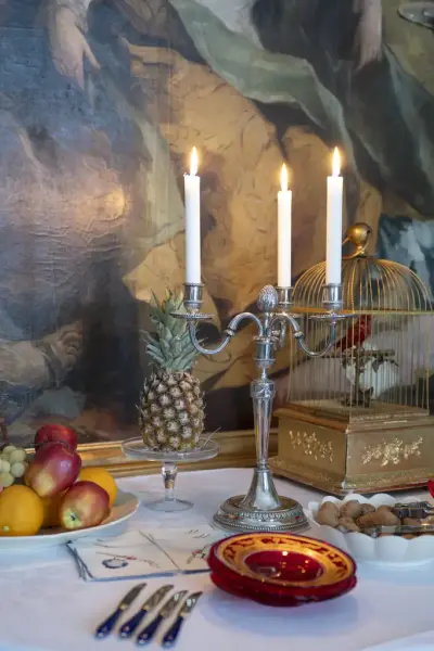 detaljer fra et bord: tente stearinlys, frukt på et fat, en fugl i bur, nøtter, kniver og tallerkener. 