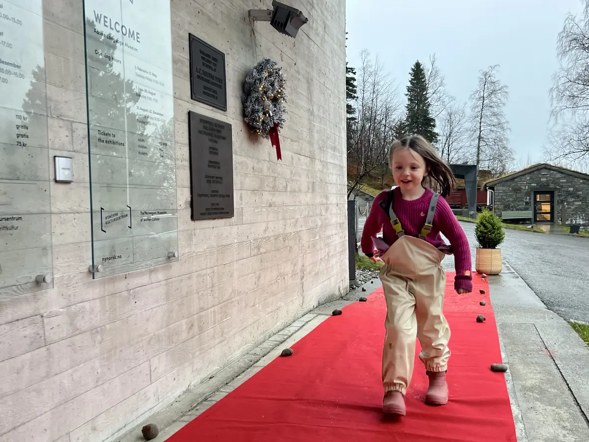 Smilande jente med regnbukse, støvlar og ullgenser kjem mot kamera på raud løpar