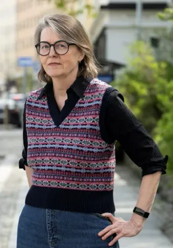 Kvinne med halvlangt hår til skuldrene. Hun har på seg olabukser, en strikkevest/genser og briller. 