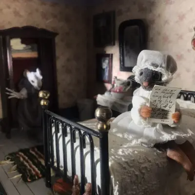 Foto av detalj fra diorama i utstillingen "Skyggesider". En mus sitter på en gammeldags metallseng og leser et brev. Musen har på seg nattlue og nattkjole. I bakgrunnen trer en skjelettaktig mus ut av et klesskap.