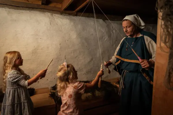 En voksen iført jernalderkjole lager vennskapsbånd i langhuset med to jenter