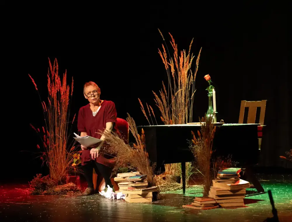 Gyldent opplyst skodespelar på ei scene med mørk bakvegg og strå, møblar og bokstablar som kulissar