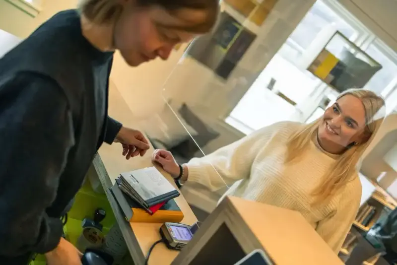 Ein tilsett hjelper ei smilande ung kvinne ved ein disk med bøker og betalingsterminal.