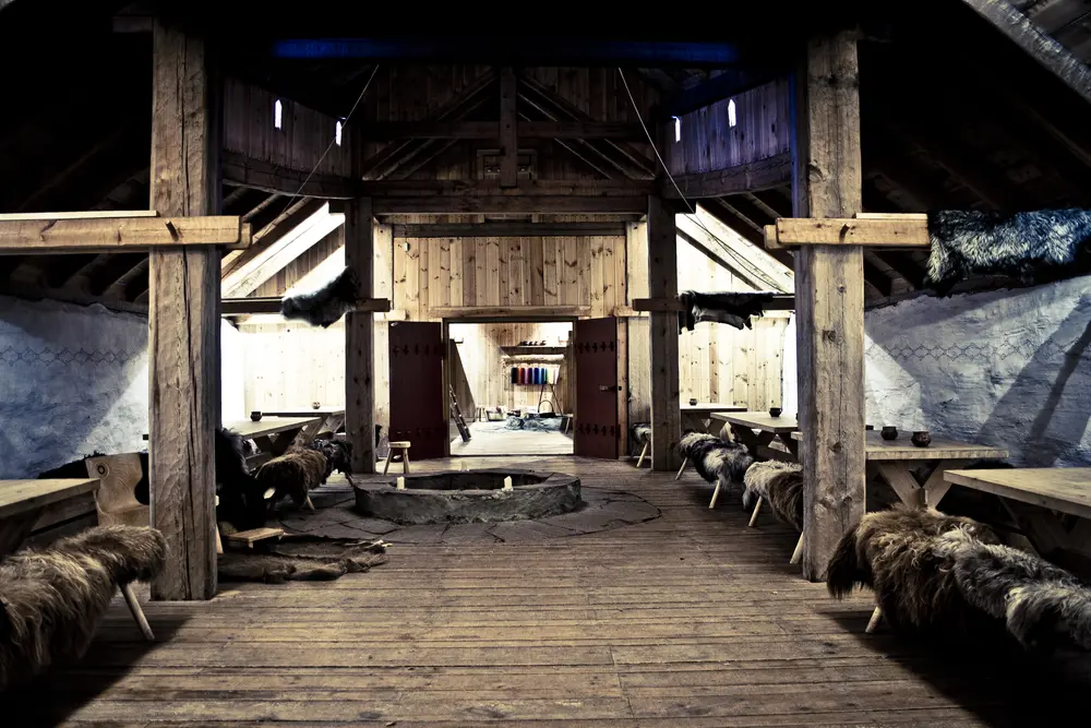 Inne i langhuset er det mørkt, det er høyt under taket og et ildsted midt på gulvet. Langs veggene er det langbord med benker rundt som har på seg saueskinn