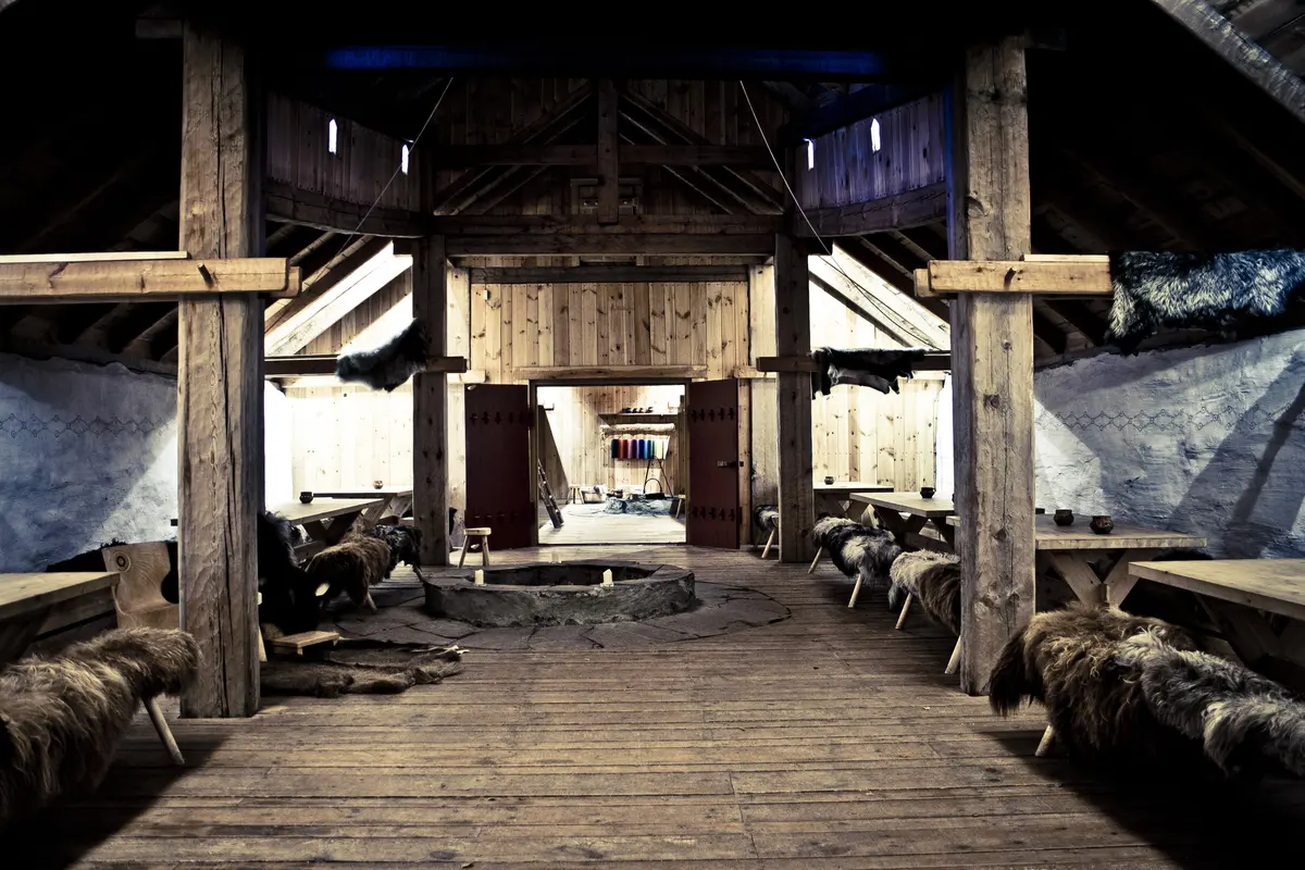 Inne i langhuset er det mørkt, det er høyt under taket og et ildsted midt på gulvet. Langs veggene er det langbord med benker rundt som har på seg saueskinn