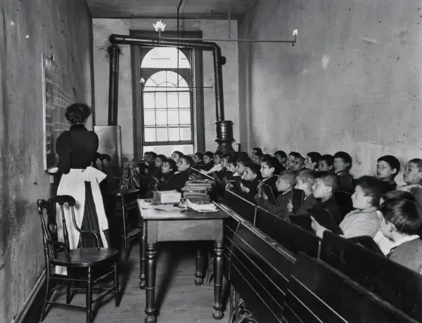 Et svart-hvitt bilde av barn som sitter tett sammen på benker i noe som kan være et trangt klasserom. En kvinnelig lærer står med ryggen til og peker på en tavle. 