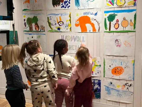 Fire barnehagebarn med ryggen til som ser og peikar på teikningar på barnekunstutstillinga. 