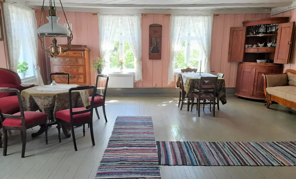 Stue med to sittegrupper, tre vinduer, chiffoniere og hjørneskap. 