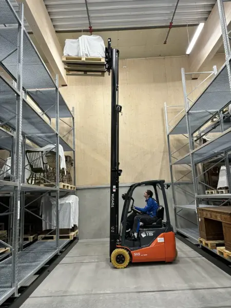 En mann i en gaffeltruck hever en pall med en gjenstand opp mot en høy hylle
