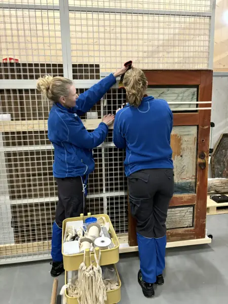 To kvinner iført blå og sort arbeidsuniform sikrer en gammel dør til en gittervegg